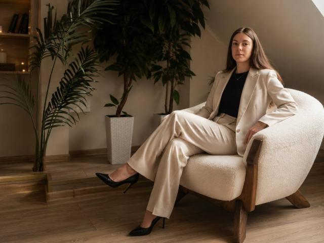 NicoleInfinity está sentada en una silla blanca, vestida con un traje color beige, una blusa negra y tacones negros, con el pelo largo y castaño, luciendo tranquila, en una habitación con suelos de madera y plantas..