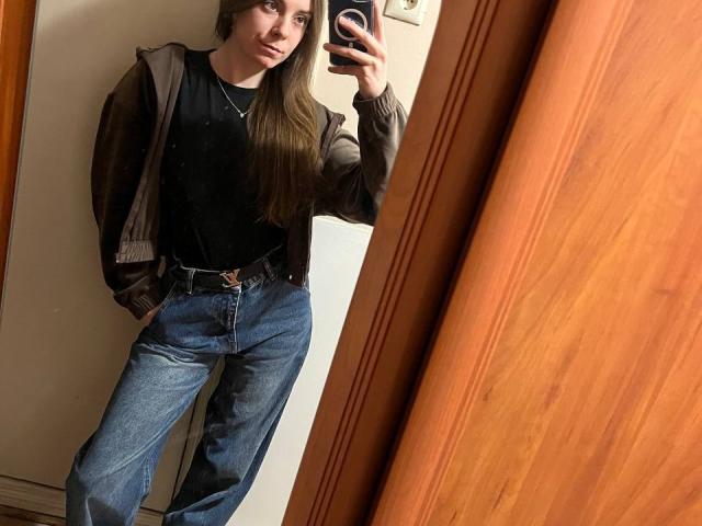NicoleInfinity, cheveux longs et bruns, chemise noire, veste marron, jean bleu, mains dans les poches, selfie devant un miroir, tenant un téléphone, porte en bois, mur blanc, style décontracté, sourire discret..