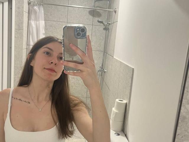 NicoleInfinity with long brown hair, white tank top, small tits, tattoo on left shoulder, holding phone, smiling in bathroom mirror selfie, toilet paper roll on right.