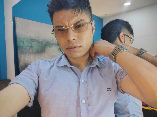 AquilesTaylor, wearing glasses and a light blue shirt, looks serious with a hand on his neck, showing a wristwatch. Reflection in the mirror shows his back.