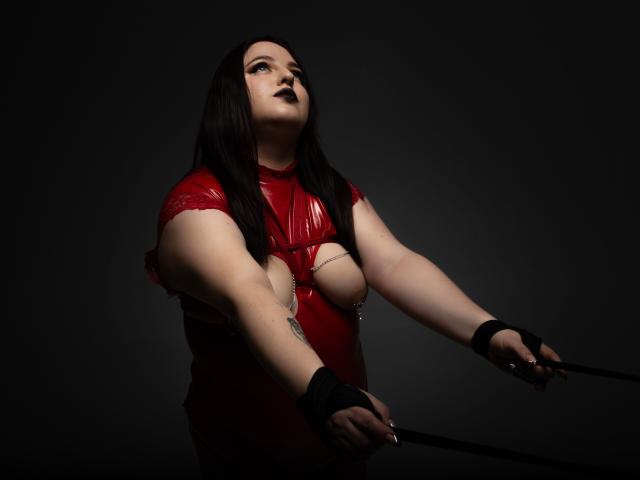 AzucenaSuency, chubby with pale skin, dark lipstick, red latex dress exposing her tits, black gloves, holding reins, dark background, looking up with a slightly open mouth.