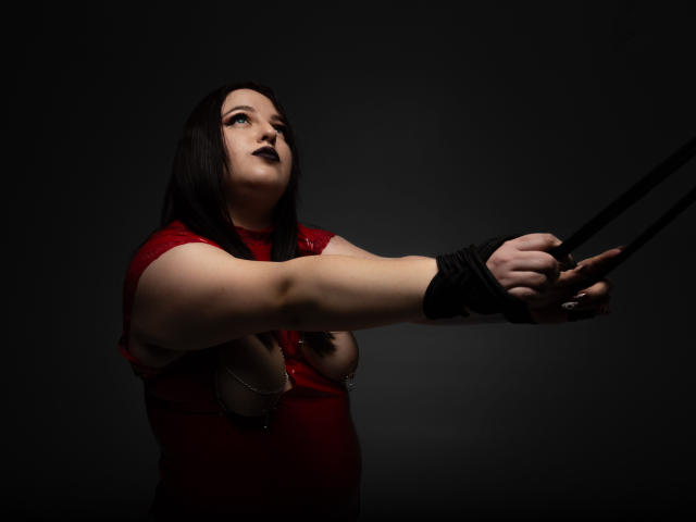 AzucenaSuency with dark hair, red top, and black wrist restraints, holding black ropes, looking up with dark lipstick, showing bare boobs with nipple chains, in a dark setting.