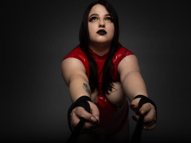 AzucenaSuency with dark lipstick and black hair, wearing a red latex top, reaching forward with black-gloved hands, chain across her chest, tattoo on her right forearm.