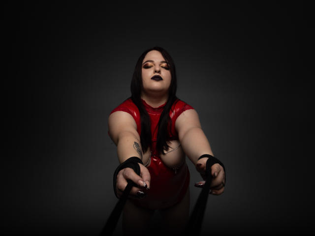 AzucenaSuency, with black lipstick, red latex, and long black hair, reaching forward with black wrist restraints, showing her tattooed arm and pale skin.