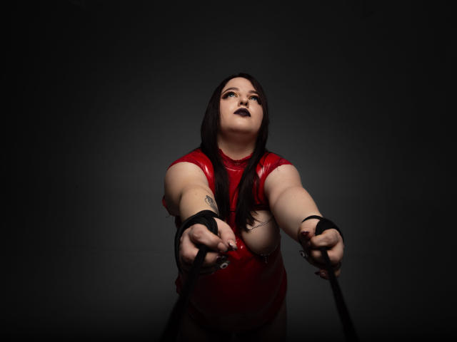 AzucenaSuency in red latex, reaching forward with black wrist cuffs, dark lipstick, long black hair, and a tattoo on her right arm, looking up.