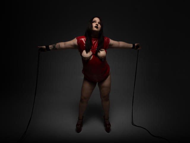 AzucenaSuency with black lipstick, red latex outfit, exposed boobs, tattoo on right arm, standing, arms out, holding black ropes, red heels, dark background, looking up, slightly open mouth.