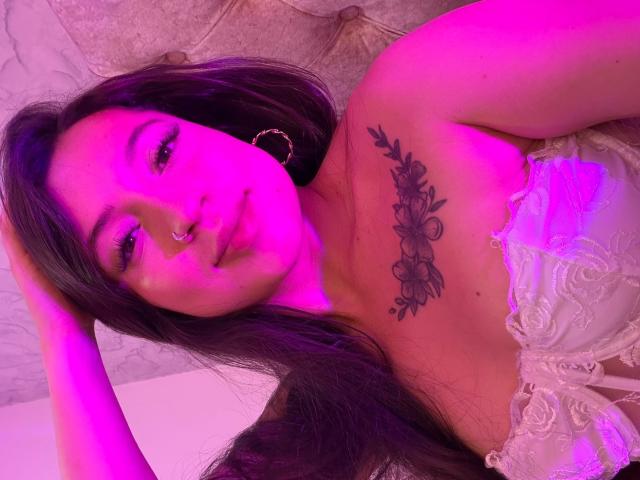 MiaAlessia, avec de longs cheveux noirs, un anneau au nez et un tatouage floral sur la poitrine, portant de la lingerie en dentelle blanche, souriante, éclairage rose et violet, une main dans les cheveux..