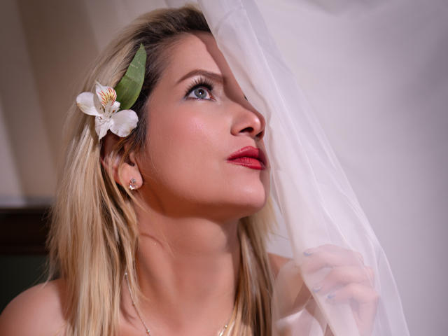 GiuliaAddams with blonde hair, blue eyes, red lipstick, white flower in hair, holding sheer curtain, looking up, wearing gold earrings, in a soft-lit, indoor setting.