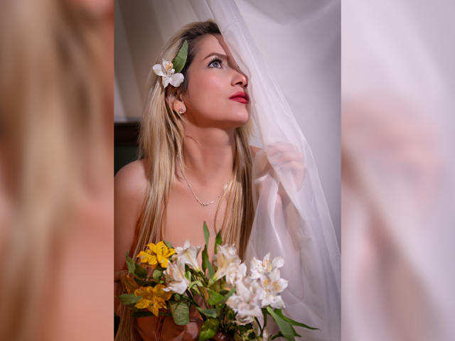 GiuliaAddams, avec de longs cheveux blonds, des fleurs blanches dans les cheveux, du rouge à lèvres, des boucles d'oreilles dorées, et un bouquet de fleurs, regardant vers le haut à travers un rideau blanc transparent..