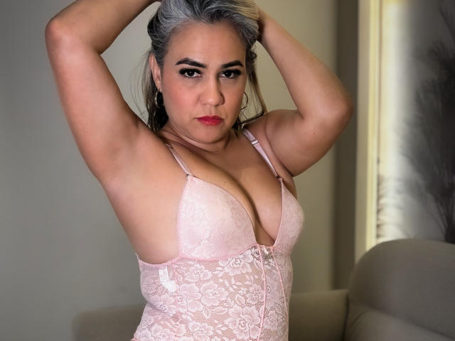 CamilaNoer, les bras levés, portant de la lingerie en dentelle rose, montrant ses seins, avec des cheveux gris, du rouge à lèvres rouge et une expression sérieuse, debout dans une pièce avec un éclairage doux..