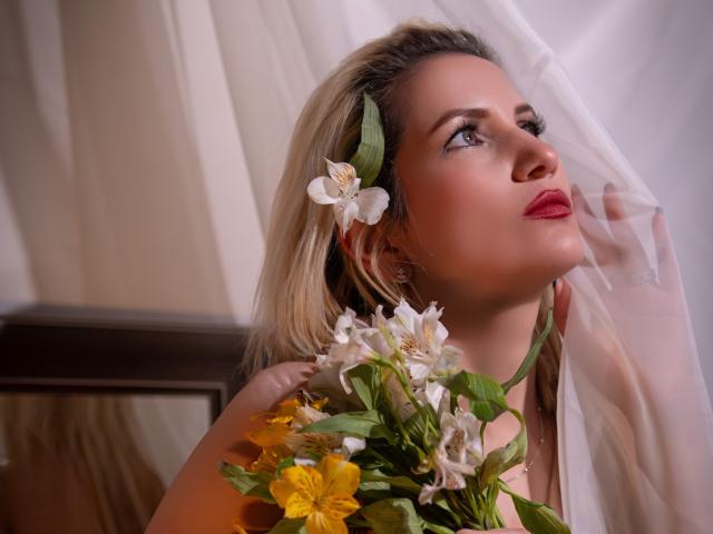  GiuliaAddams with blonde hair, white flowers in her hair, green leaf, and yellow flowers, looking up with red lipstick, holding sheer curtain, soft lighting, focused expression.
