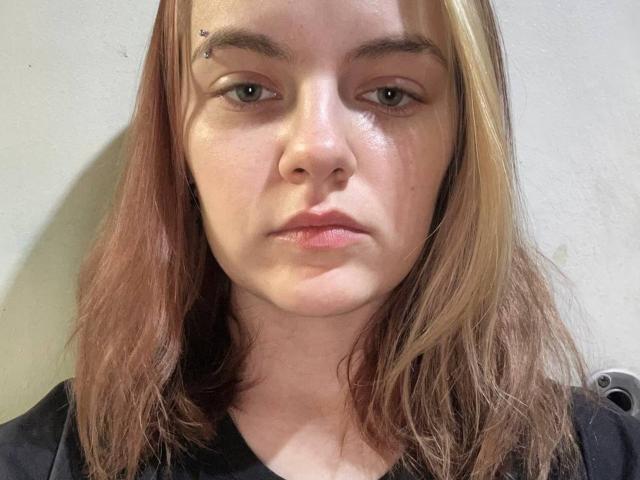 Nena Selic a les cheveux châtain clair, mi-longs, porte un haut noir et a une expression neutre, avec de petits clous métalliques sur son sourcil droit. Sa peau est légèrement brillante..