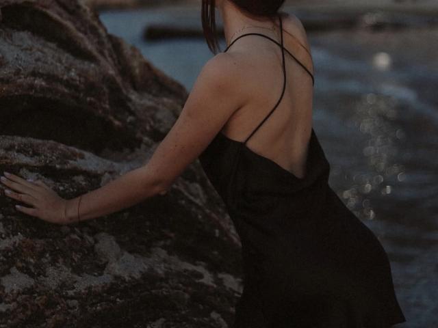 Nikoletes with her back to the camera, wearing a black, spaghetti-strap dress, leaning against a rock by the water. Her ass is slightly visible. Dark, wavy hair.