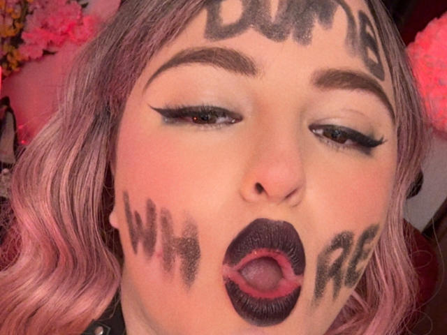 AzucenaSuency's face with pink hair, black eyebrows, and dark lipstick, has black paint on forehead, cheeks, and lips. Her mouth is open, showing a pink tongue.