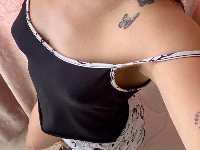 SophiaMarx's black tank top with white straps, showing her small tits and butterfly tattoo on her chest, wearing white print pants, pink curtain in background.