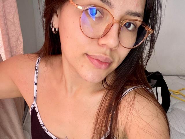 SophiaMarx with brown hair, wearing tortoiseshell glasses, has small nose and light pink lips, wearing a black and white patterned top, showing a slight smile, and a small nose piercing.