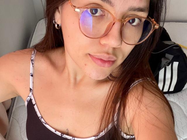 SophiaMarx with brown hair, wearing glasses and a black camisole with white pattern, has a cute face, light skin, and small tits, looking at the camera.