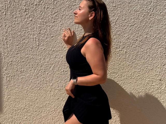 Mira-Moon-hot hat gebräunte Haut, braunes Haar und trägt ein schwarzes, ärmelloses Kleid mit einer Uhr. Sie steht vor einer strukturierten weißen Wand, blickt zur Seite und hat die Hände erhoben..