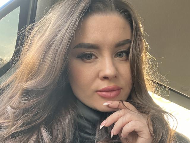 OlesyaMay a de longs cheveux bruns ondulés, de pleines lèvres roses, et elle porte un col roulé noir. Elle regarde l'appareil photo et touche son menton avec son doigt..