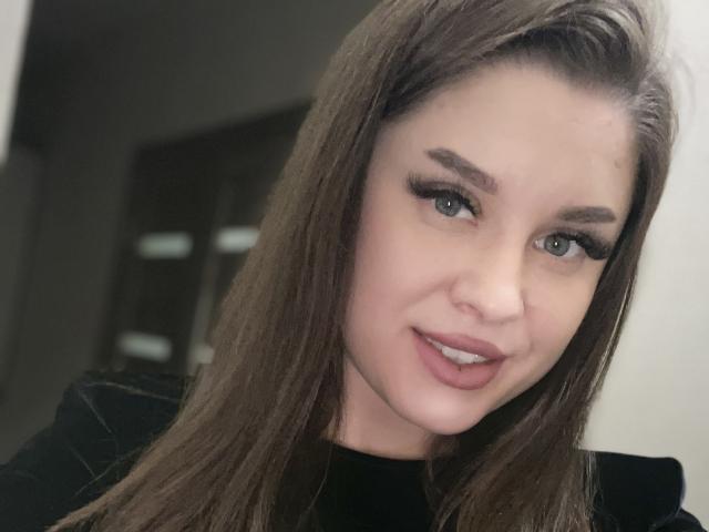 OlesyaMay a de longs cheveux bruns, des yeux verts et des lèvres pulpeuses avec du rouge à lèvres rose. Elle porte des vêtements noirs et regarde directement l'appareil photo avec un léger sourire..