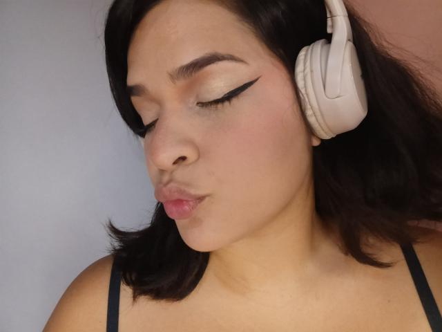AntonelaPark with puffy pink lips, black winged eyeliner, and closed eyes, wearing white headphones, black strappy top, and short dark hair, looking down.