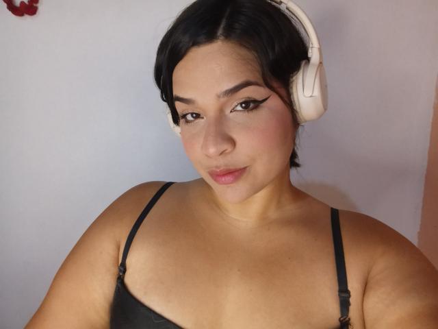 AntonelaPark with dark hair, wearing white headphones, black bra, and winged eyeliner, has a cute, slightly chubby face, small tits, and a subtle smile.