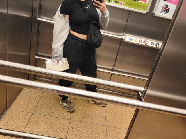 Leonora, habillée d'un crop top noir, d'un pantalon noir, d'une veste grise, tenant un téléphone, un sac noir, une boîte blanche, des chaussures argentées, debout dans un ascenseur en métal, prend un selfie devant un miroir..