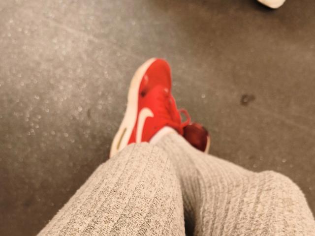 Leonora-hots roter Sneaker auf einem strukturierten, grauen Teppich, wobei ihre grauen, gerippten Hosen zu sehen sind. Der Sneaker hat weiße Streifen, und Leonora-hots Bein ist leicht gebeugt..