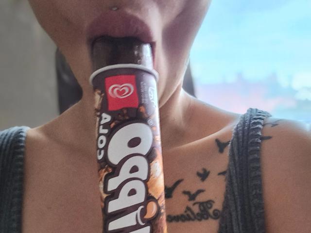 Leonora-hot hat ihre Lippen um einen Schokoladen-Pop Rocks-Stick, zeigt ein Nasenpiercing und hat ein Tattoo von Vögeln auf ihrer Brust. Sie trägt ein graues, geripptes Tanktop..
