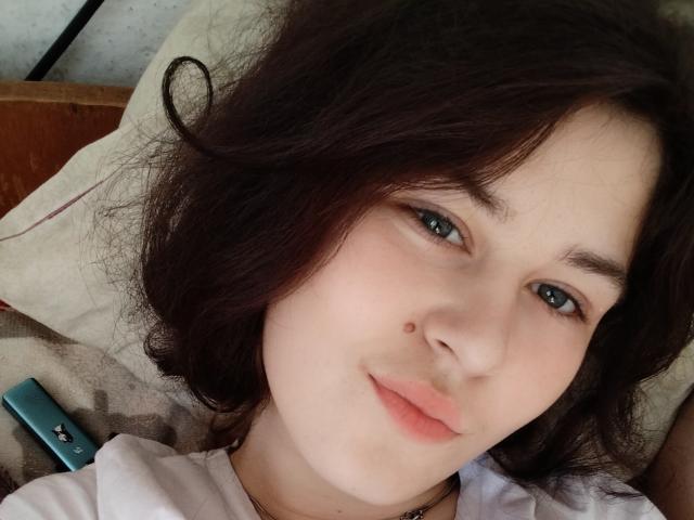 Tory Tayler tem pele clara, cabelo escuro e ondulado, olhos azuis e uma pequena marca de beleza na bochecha direita. Ela está vestindo uma camisa branca, deitada, com um controle remoto azul..