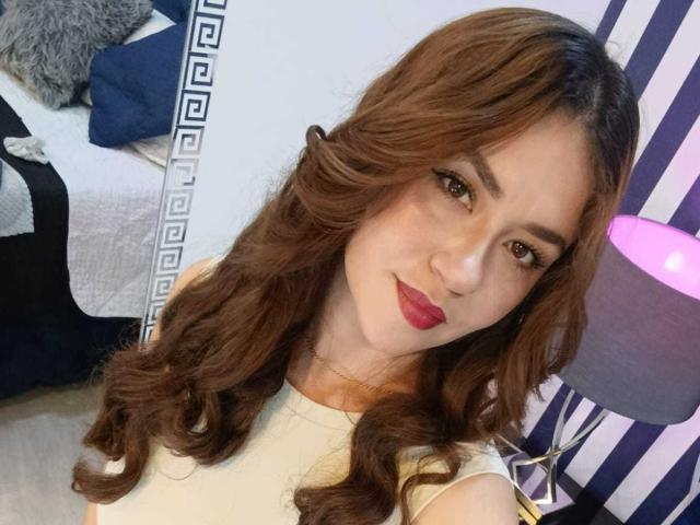 Theresa Larson tem cabelos castanhos ondulados, batom vermelho e olhos castanhos, vestindo uma camisa branca, com um abajur roxo ao fundo. Seus seios não estão visíveis..