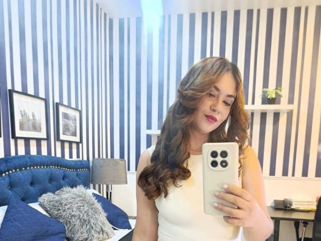 Theresa Larson, met golvend bruin haar, rode lippenstift, een wit mouwloos topje, die een iPhone vasthoudt en een selfie maakt in een kamer met blauwe strepen. Op de achtergrond staat een bed met een blauw gestoffeerd hoofdeinde en een grijs kussen..