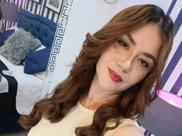 TheresaLarson hat welliges, braunes Haar, roten Lippenstift und ein süßes Lächeln. Sie trägt ein beiges Oberteil, und ihre Brüste sind teilweise sichtbar. Sie macht ein Selfie..