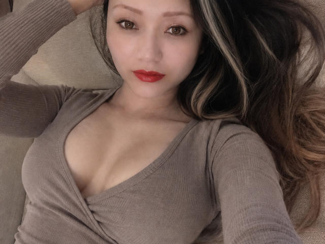 VioletG, con cabello largo y oscuro, lápiz labial rojo y una blusa beige de punto con escote pronunciado que deja ver sus senos, está acostada en un sofá y mirando a la cámara..