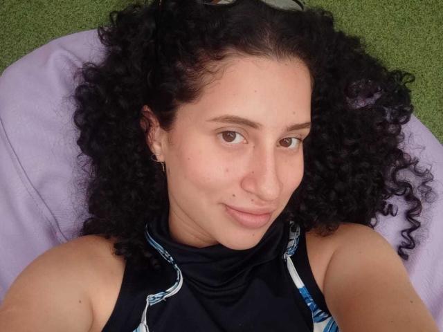 NoaxxMalak, avec des cheveux noirs bouclés, une peau brun clair, un petit nez, un sourire discret, portant un haut sombre sans manches, allongée sur un oreiller violet, fond verte..