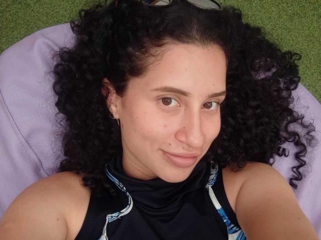 NoaxxMalak tiene el pelo negro y rizado, ojos marrones claros y una ligera sonrisa. Lleva una camiseta oscura sin mangas, está acostada sobre un cojín morado, y en el fondo se ve hierba verde..