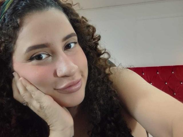 NoaxxMalak, com cabelos castanhos encaracolados, pele clara e olhos castanhos, apoiando o rosto na mão e sorrindo suavemente. Ela está usando uma maquiagem discreta e uma alça preta..