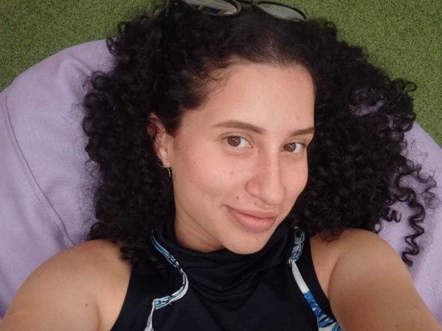 NoaxxMalak, com cabelo preto encaracolado, pele clara, nariz pequeno e um sorriso discreto, está deitada em um travesseiro roxo, vestindo uma blusa preta sem mangas, e usa pequenos brincos e óculos na cabeça..