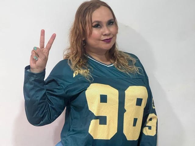 MiaCapricce with wavy blonde hair, wearing a teal football jersey with gold numbers, flashing a peace sign with her right hand, showing off her tits and green ring.