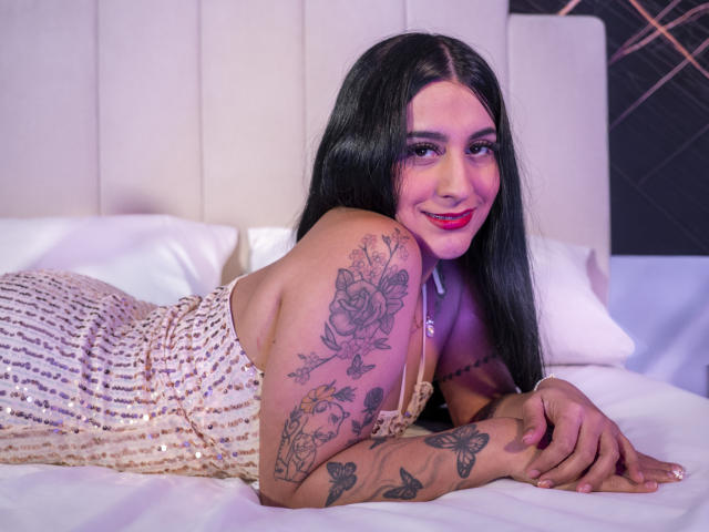 SamyRush, con lunghi capelli neri, rossetto rosa e tatuaggi sulle braccia e sul petto, sdraiata sul letto, indossa un abito dorato scintillante, sorride alla fotocamera, con le mani appoggiate sul letto bianco..