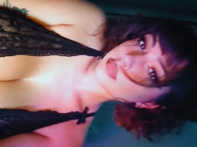 NaughtyMia, atractiva con cabello castaño rizado, vistiendo lencería negra de encaje, mostrando sus pechos, con una expresión juguetona y ligeramente traviesa, y maquillaje de ojos oscuro y ahumado en una foto con poca luz..