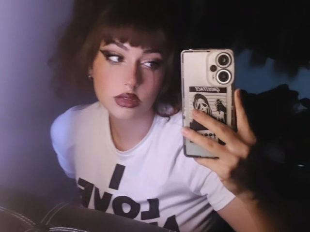 NaughtyMia-hot with dark hair, thick eyebrows, and purple lipstick, wearing a white shirt, taking a selfie with a phone in a dimly lit room, showing her pouty face.
