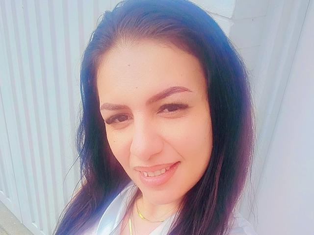 CamilleRiver has dark, wavy hair, light skin, and a small smile showing a single front tooth. She's wearing a white shirt with a yellow necklace.