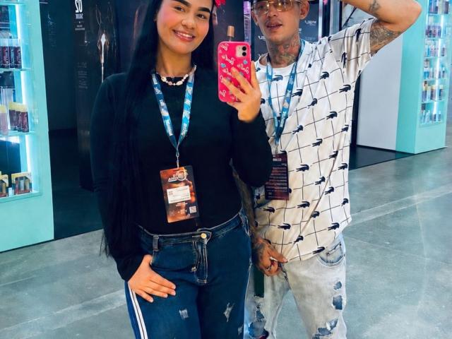 MiaaWhithe with long black hair, black top, and ripped jeans stands next to a tattooed guy in a patterned shirt, both wearing lanyards, taking a selfie with a pink phone.