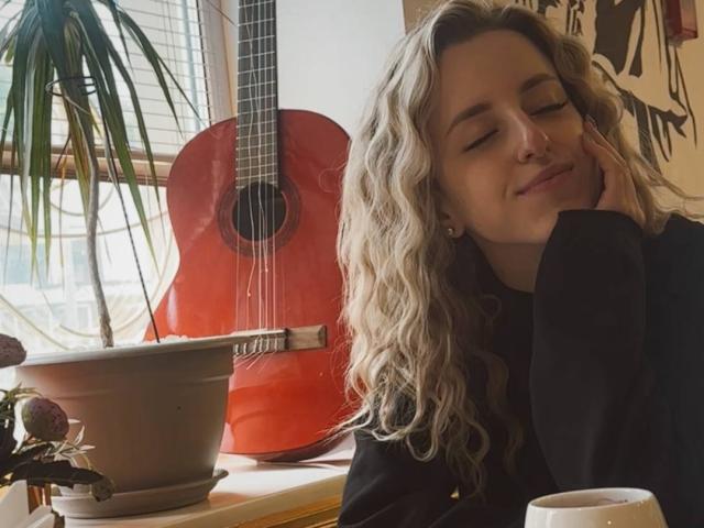 Maximina Mayhood mit welligem, blondem Haar, mit geschlossenen Augen, in einem schwarzen Oberteil, mit dem Kinn auf der Hand, einer roten Akustikgitarre, einer Topfpflanze und einer weißen Tasse im Vordergrund, lächelt sanft..