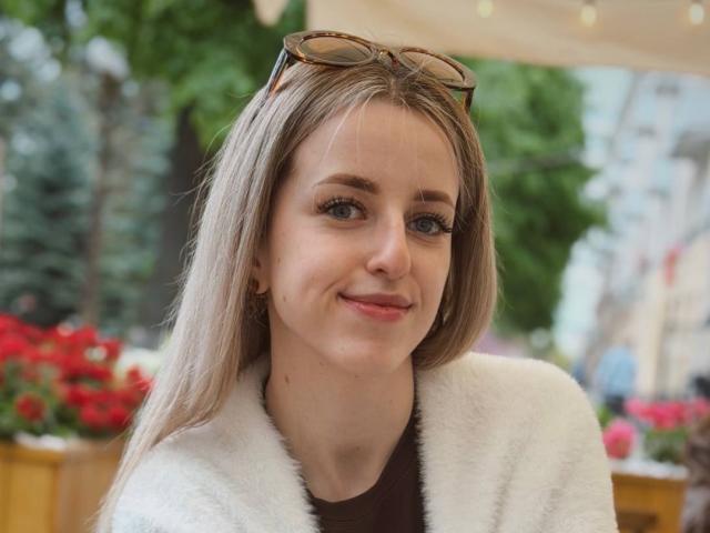 MaximinaMayhood tem cabelo loiro, olhos azuis e um sorriso adorável. Ela está vestindo um casaco branco fofo com uma parte superior marrom e óculos na cabeça. Ela é uma garota muito bonita..