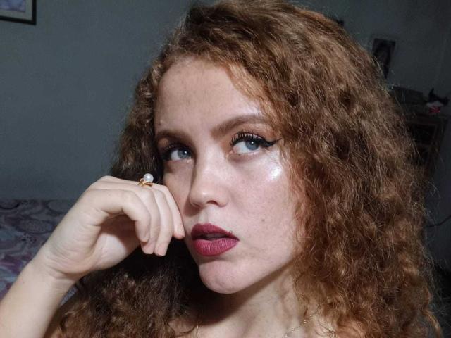 MiaSweetty69 with curly red hair, blue eyes, shiny skin, dark red lipstick, and a ring on her finger, holding her chin with a pensive look.