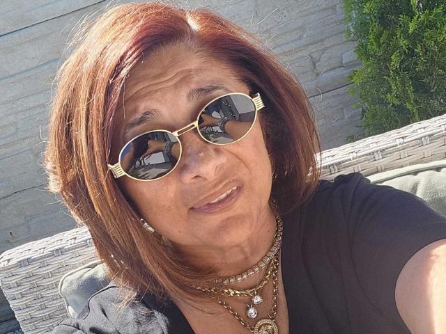 Elegante ma audace, con capelli rosso-marrone, indossa occhiali da sole rotondi dorati e diverse collane, sorride alla fotocamera, mostrando un viso segnato dalle rughe e indossando una maglietta nera..