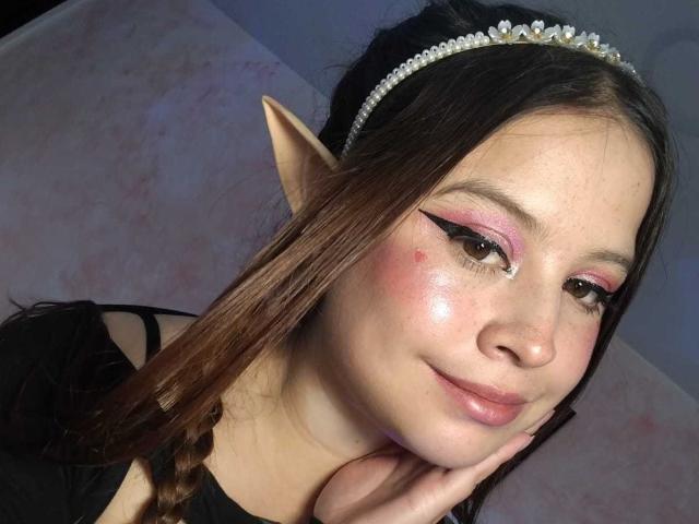 Melani Becker, met elfenoorjes, roze eyeliner en een diamanten hoofdband, heeft een kleine rode puist op haar wang, bruine ogen en een subtiele glimlach, en draagt zwart..