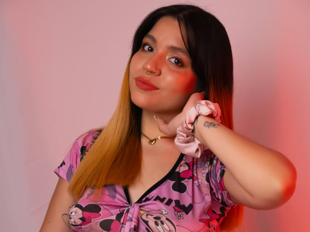 Abby Junes con cabello degradado de rubio a negro, lápiz labial rojo, una goma rosa en la muñeca, vistiendo una camiseta rosa de Mickey Mouse, un tatuaje pequeño en la muñeca, un collar dorado, una ligera sonrisa, iluminación rosa-roja..